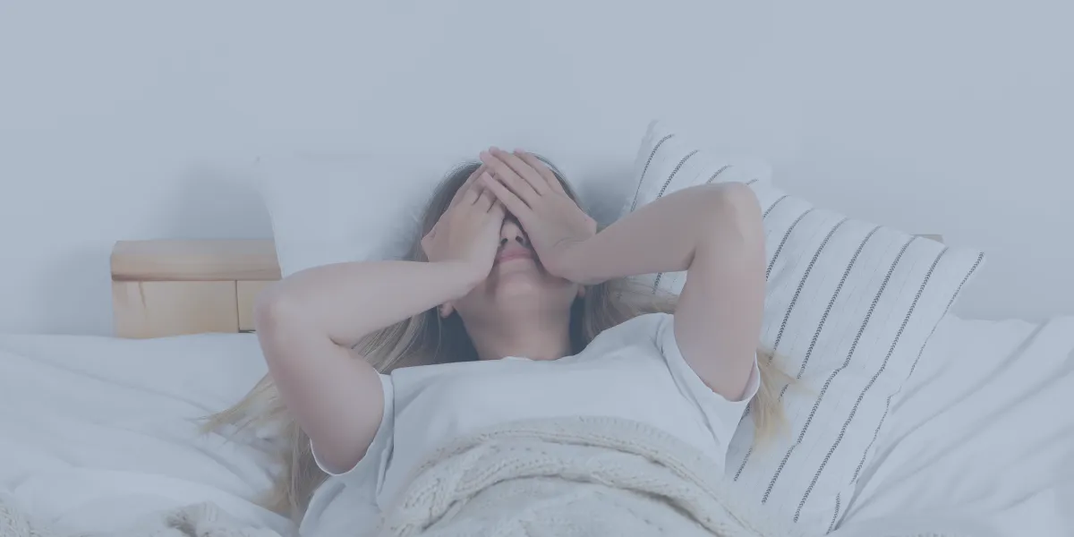 Woman lying in bed covering her face with her hands, appearing distressed