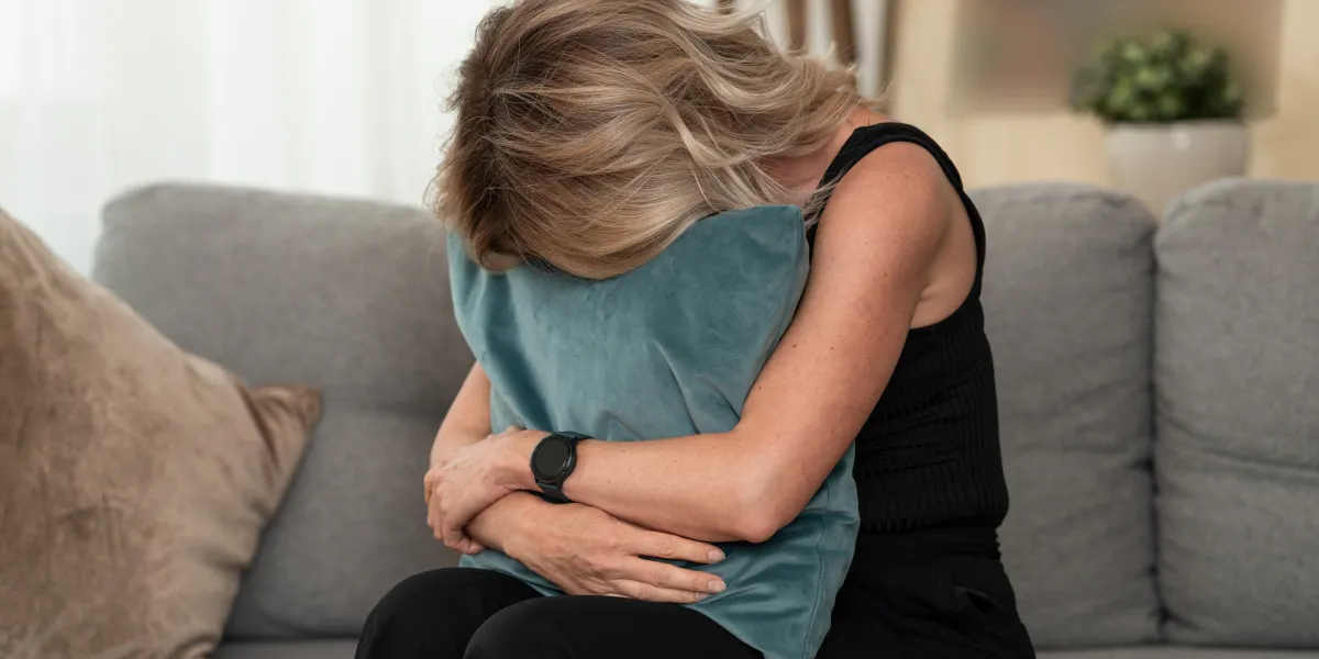 Woman hugging a pillow while sitting on a grey couch, head down in sadness